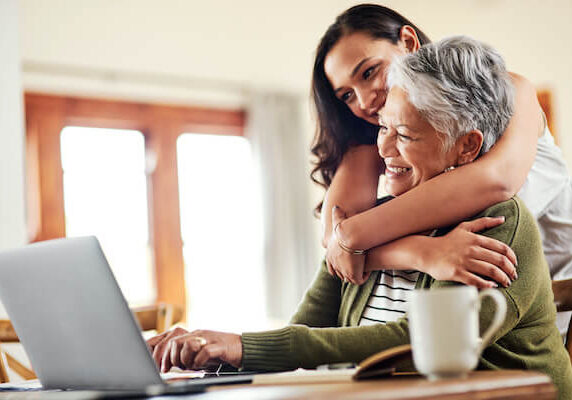A smiling older woman sits at a laptop in a sunny room, with her adult daughter hugging her.