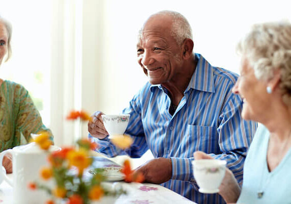 Three smiling older people sit around a table together drinking from white porcelain teacups.